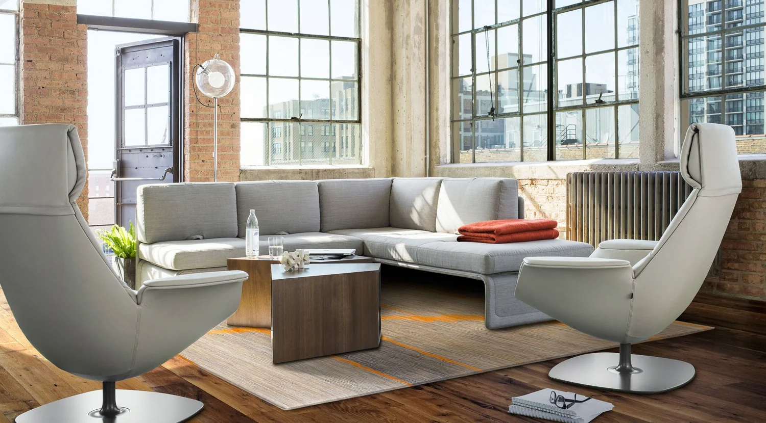 Moderne loungespace met grijze designmeubels, grote industriële ramen en een houten vloer.