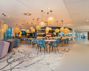 Modern café-interieur met blauwe stoelen, houten tafels en hangende rieten lampen in een lichte, open ruimte.