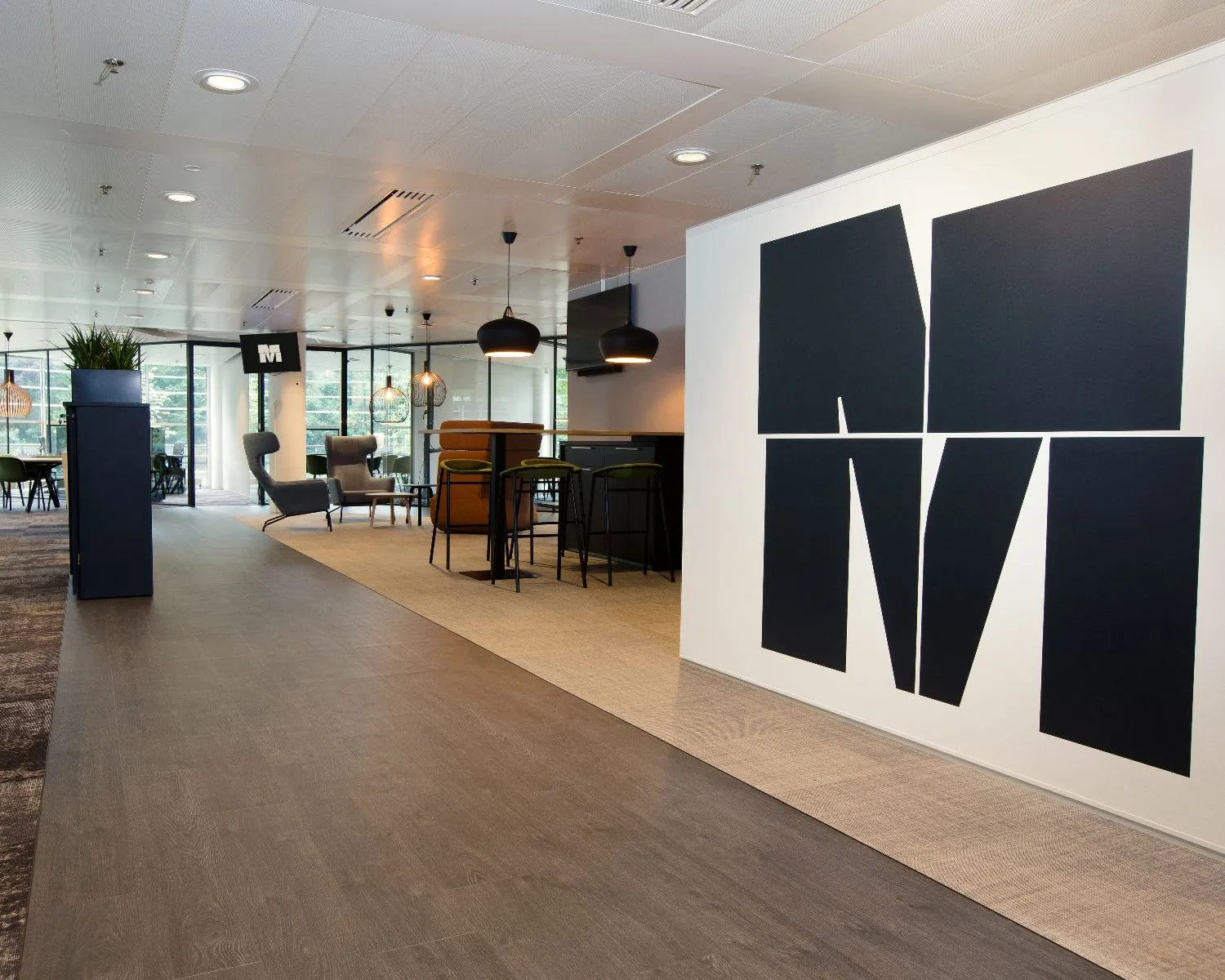 Modern kantoorinterieur met zitgedeelte, hoge tafels en een groot zwart-wit M-logo op de muur.