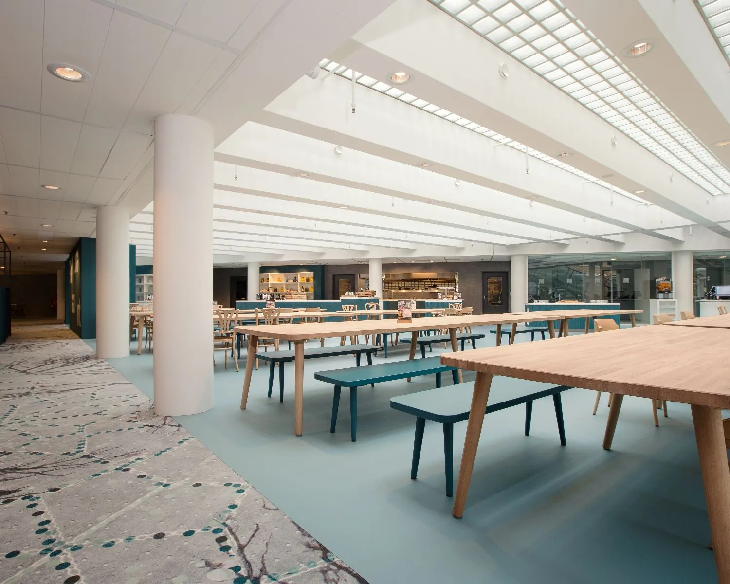 Licht en modern ingericht bedrijfsrestaurant met houten tafels en blauwe banken.