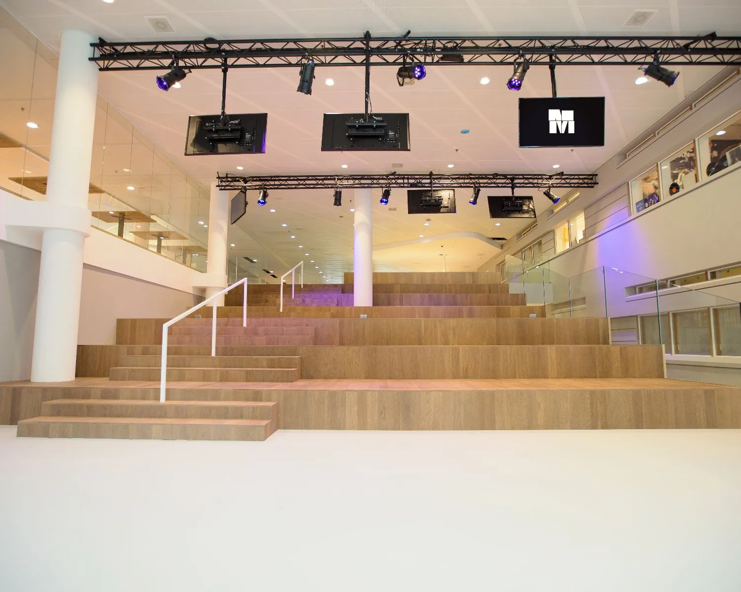 Houten tribunetrap met witte leuningen in een moderne, open binnenruimte met glazen balustrades en hangende schermen.