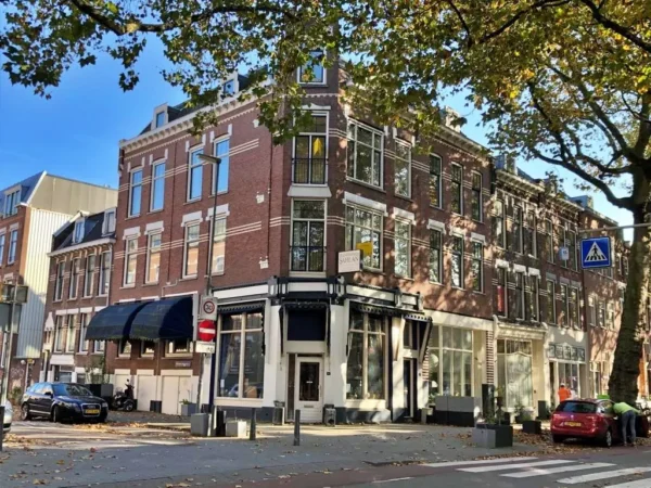Street corner in Rotterdam-Noord with historic brick buildings, trees, and parked cars on a sunny day.