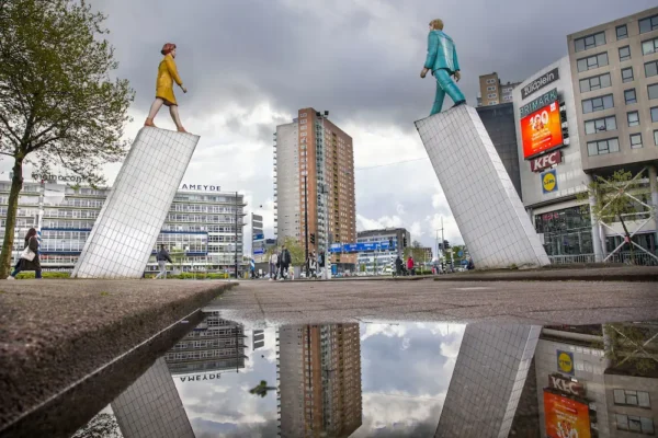 Two large statues of a man and woman face each other atop tilted pillars in a busy urban square in Rotterdam-Zuid.