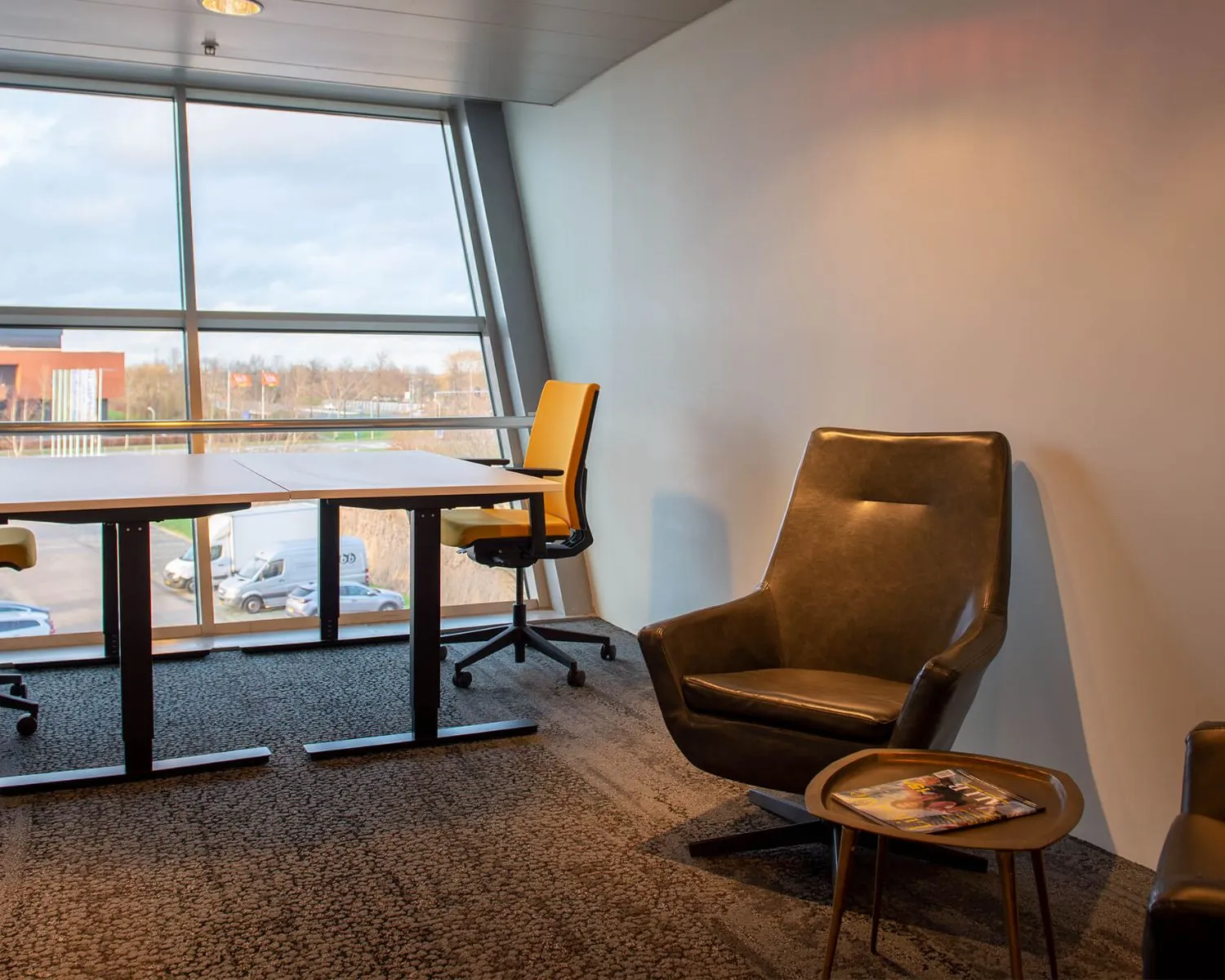 Moderne kantoorruimte met gele bureaustoelen, een leren fauteuil en een bijzettafel met tijdschriften bij een groot raam.