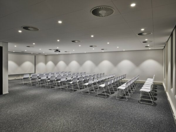 Vergaderruimte aan de Van Deventerlaan 31-51 met rijen witte stoelen, grijze vloerbedekking en een groot projectiescherm.