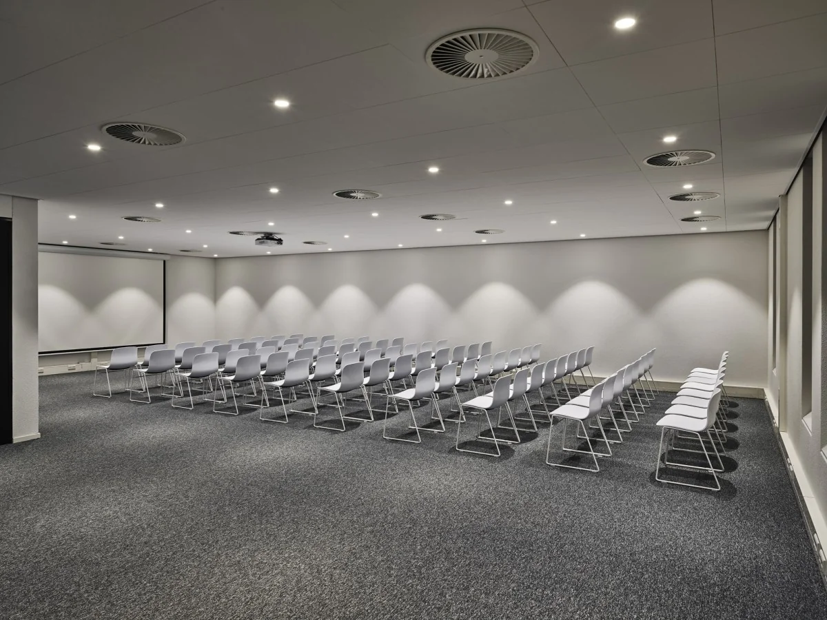 Vergaderruimte aan de Van Deventerlaan 31-51 met rijen witte stoelen, grijze vloerbedekking en een groot projectiescherm.