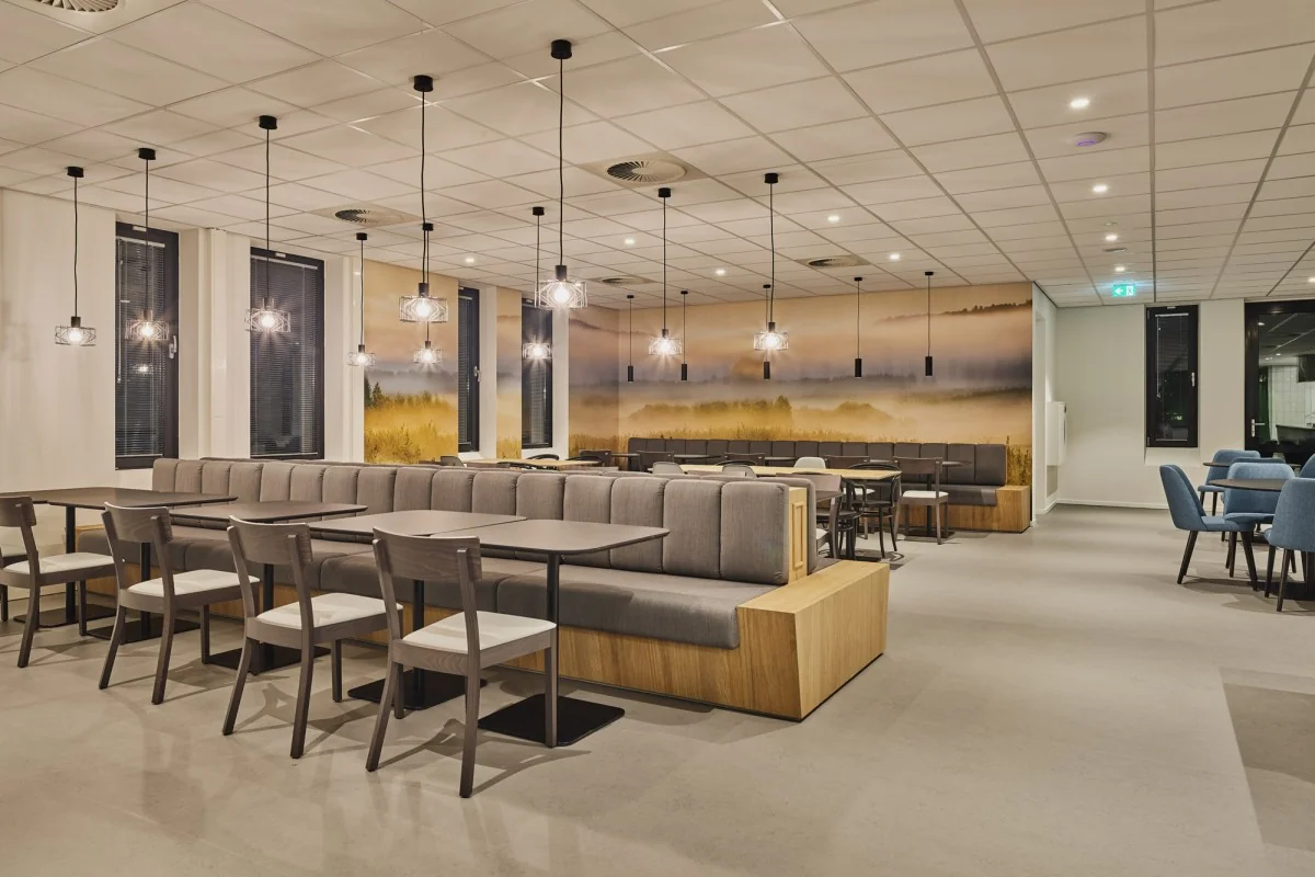 Moderne bedrijfsrestaurant aan de Van Deventerlaan met sfeervolle verlichting, wandbanken en natuurprint op de muur.
