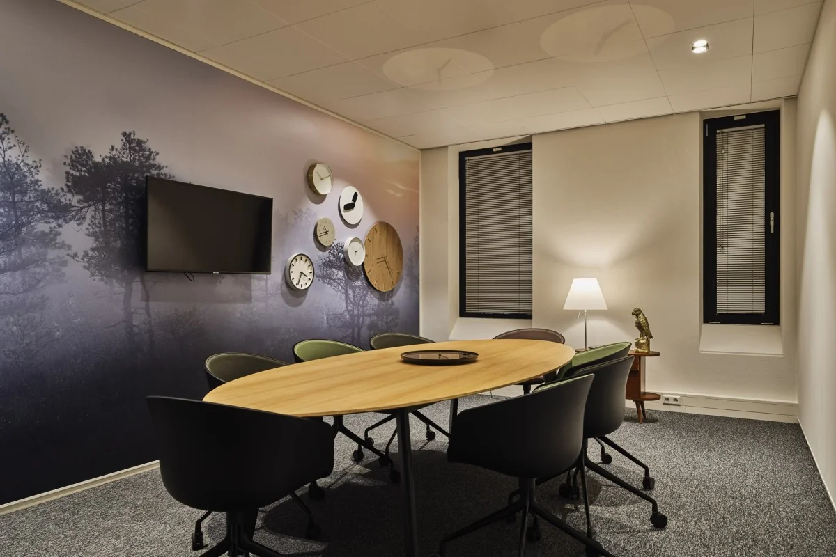 Vergaderruimte aan de Van Deventerlaan 31-51 met ovale houten tafel, zes stoelen, wand met klokken en tv, en een staande lamp bij het raam.