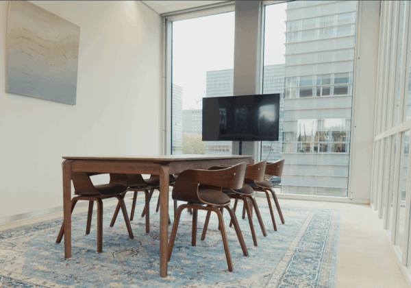 Vergaderkamer met houten tafel en stoelen, een vloerkleed, een flatscreen en grote ramen met uitzicht op moderne kantoorgebouwen.