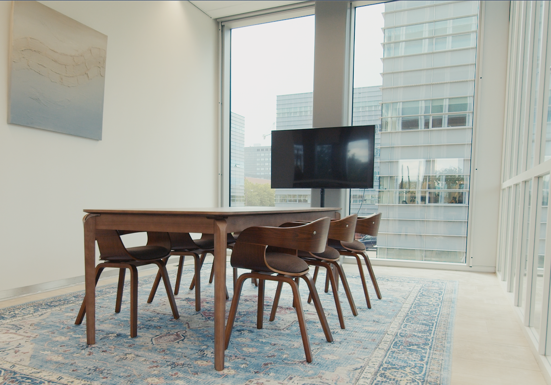 Vergaderkamer met houten tafel en stoelen, een vloerkleed, een flatscreen en grote ramen met uitzicht op moderne kantoorgebouwen.