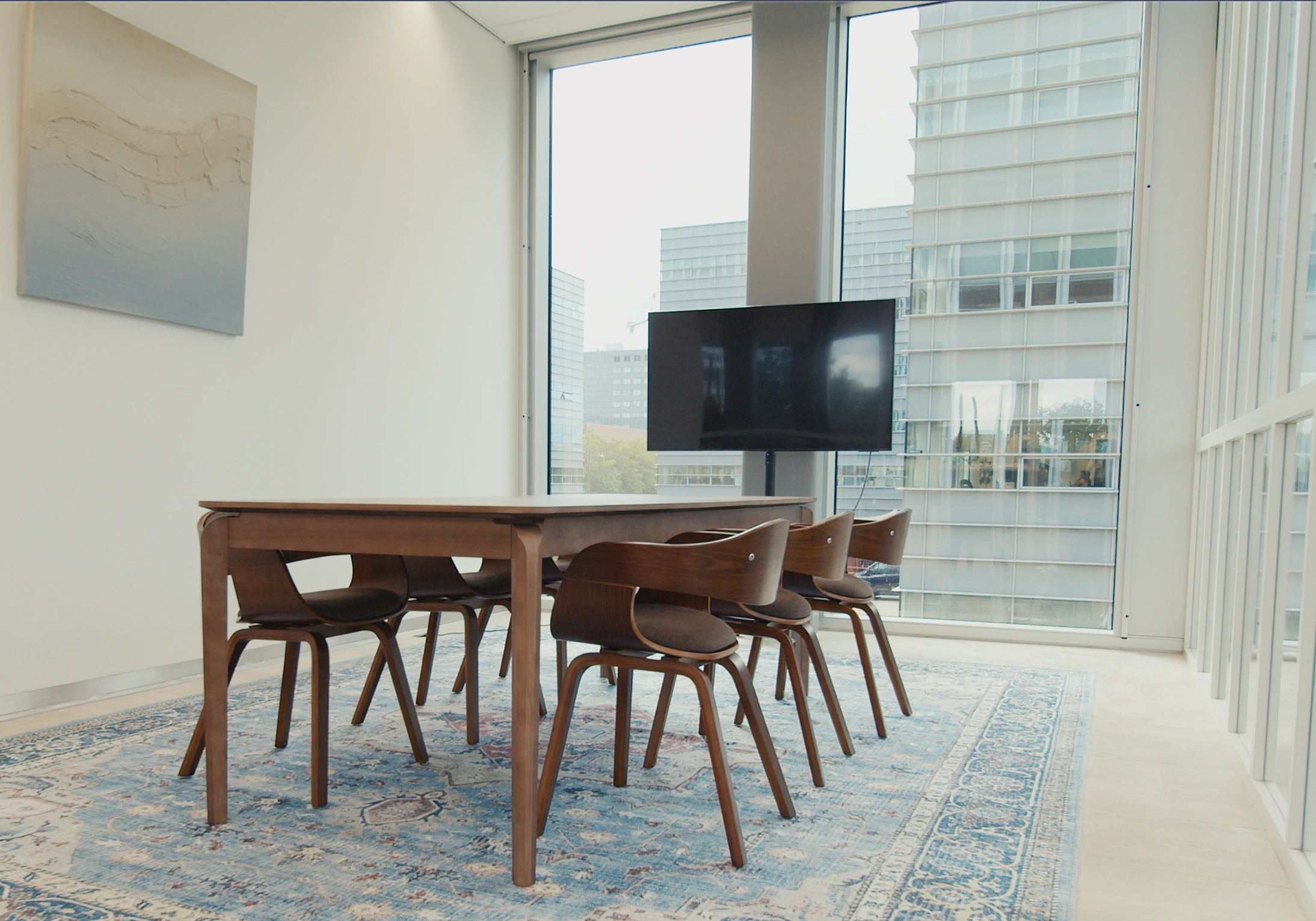 Vergaderkamer met houten tafel en stoelen, een vloerkleed, een flatscreen en grote ramen met uitzicht op moderne kantoorgebouwen.