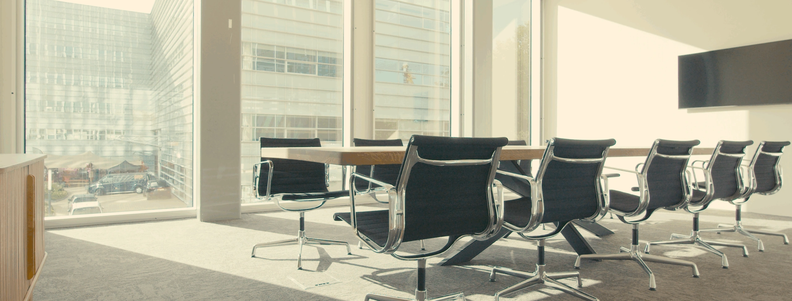 Moderne vergaderkamer met een lange houten tafel, zwarte bureaustoelen en grote ramen met uitzicht op een kantoorgebouw.