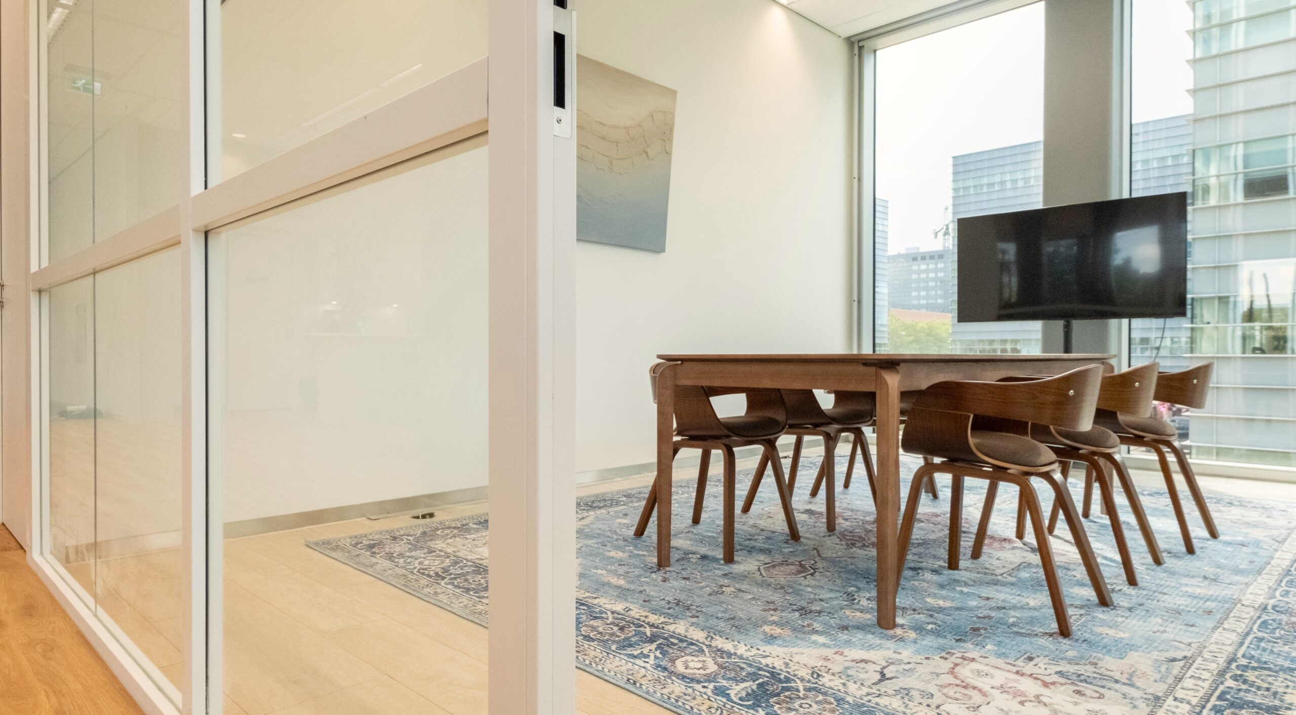 Vergaderkamer met houten tafel en stoelen, groot vloerkleed, flatscreen en glazen wanden.