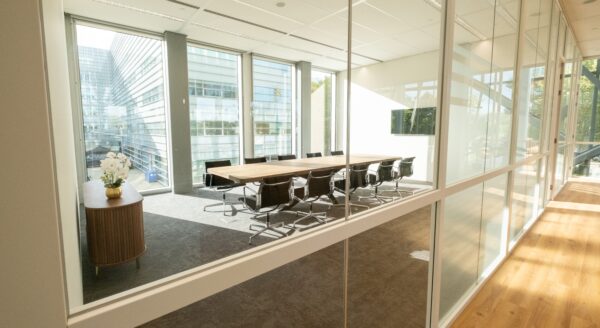 Heldere vergaderruimte met lange houten tafel, moderne stoelen en grote ramen met uitzicht op kantoorgebouwen.