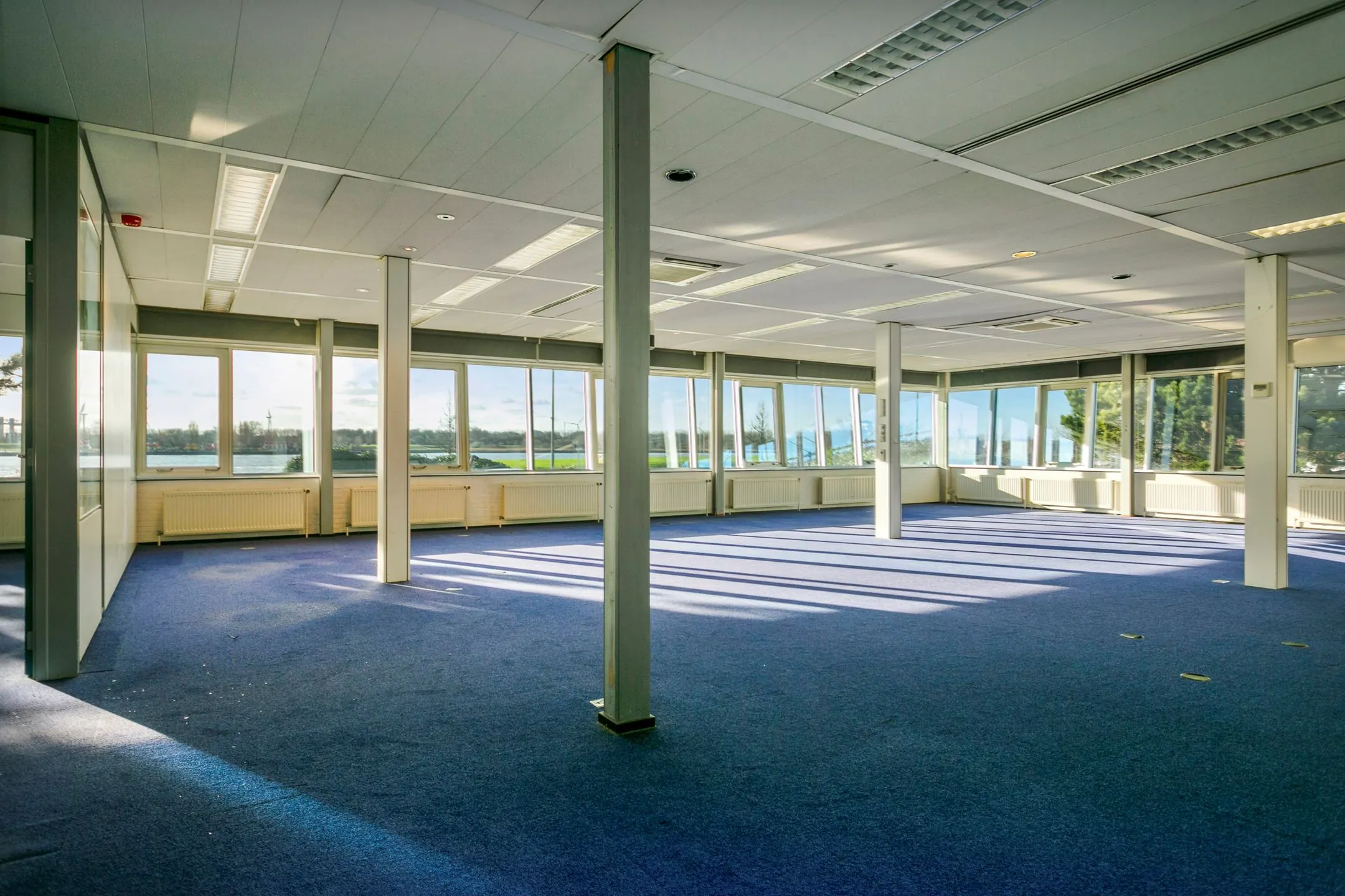 Lichte, leegstaande kantoorruimte met blauw tapijt en uitzicht op het water in Maassluis.