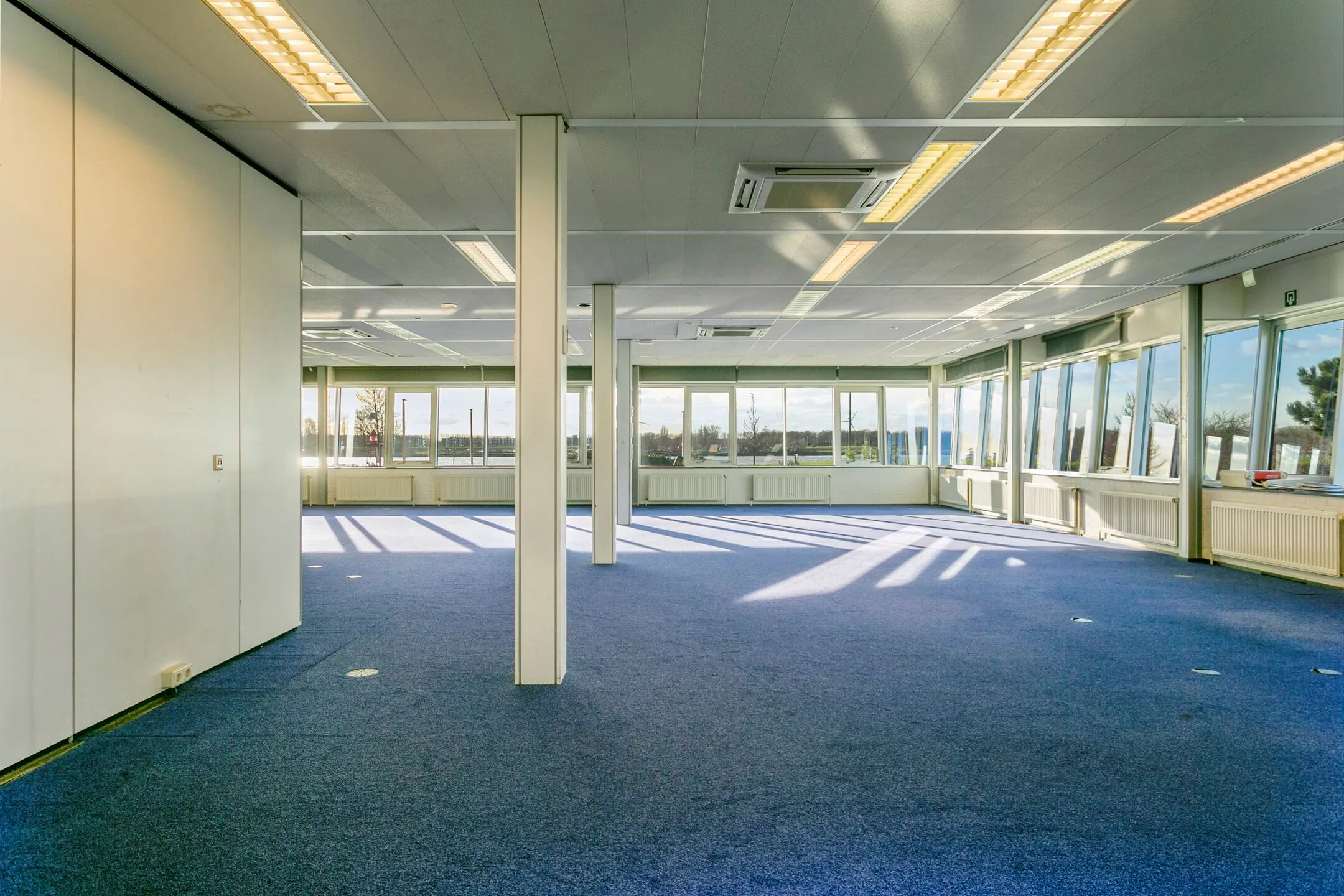 Licht en ruim kantoorpand in Maassluis met blauwe vloerbedekking en grote ramen rondom.