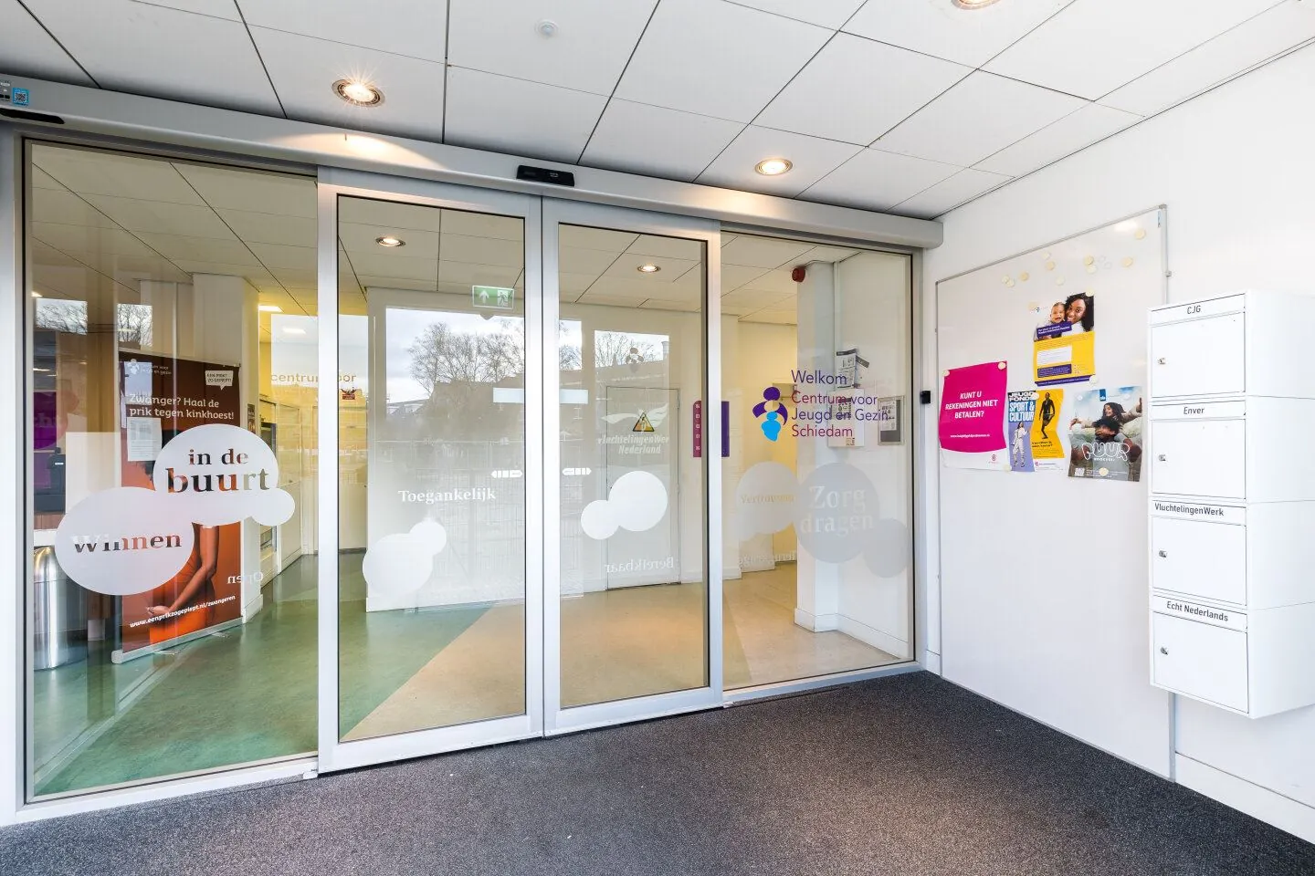 Ingang van het Centrum voor Jeugd en Gezin Schiedam met glazen schuifdeuren en informatieve posters aan de muur.