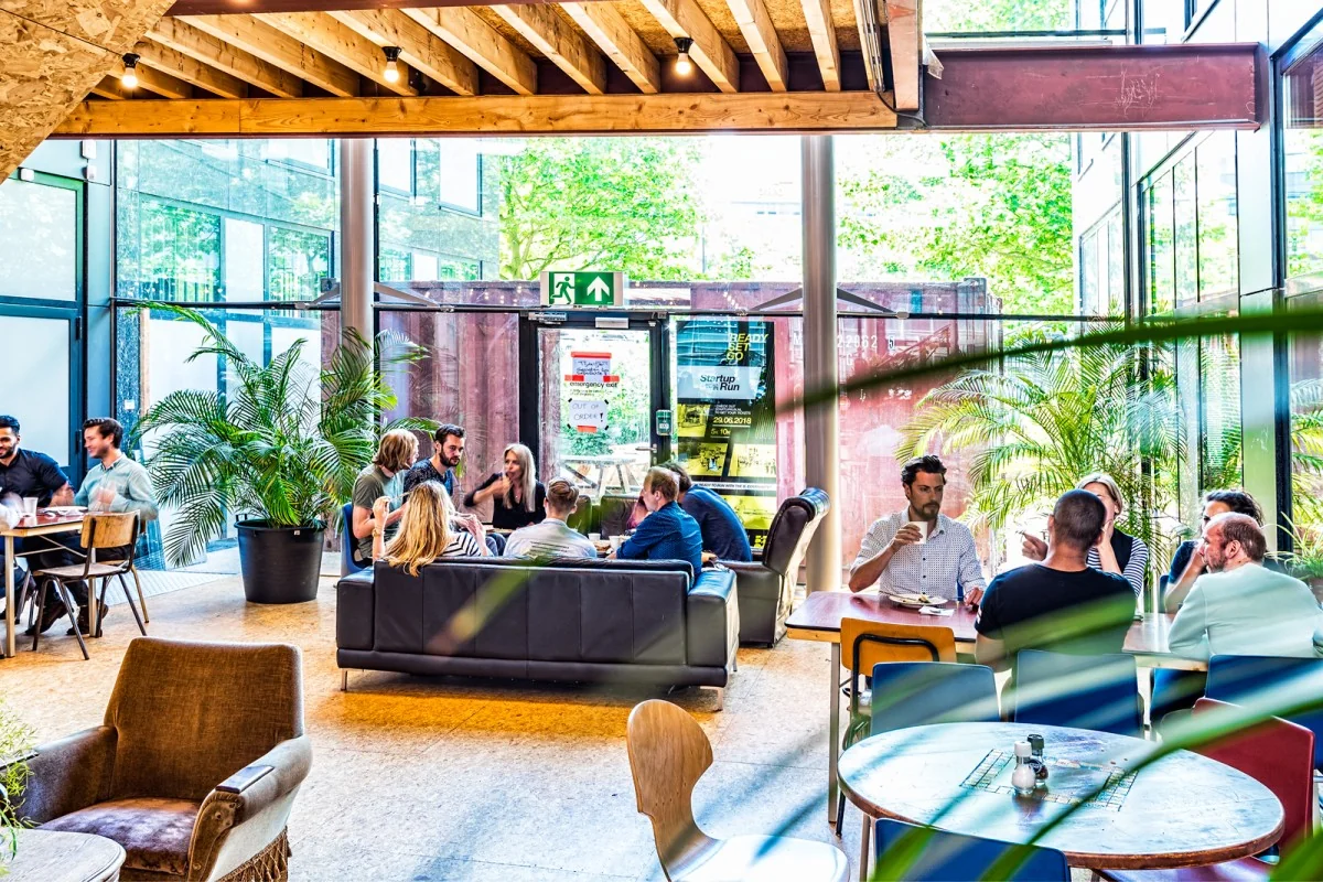Gezellige en moderne loungeruimte in een kantoorpand aan het John M. Keynesplein in Amsterdam met mensen in gesprek en omringd door grote ramen en planten.