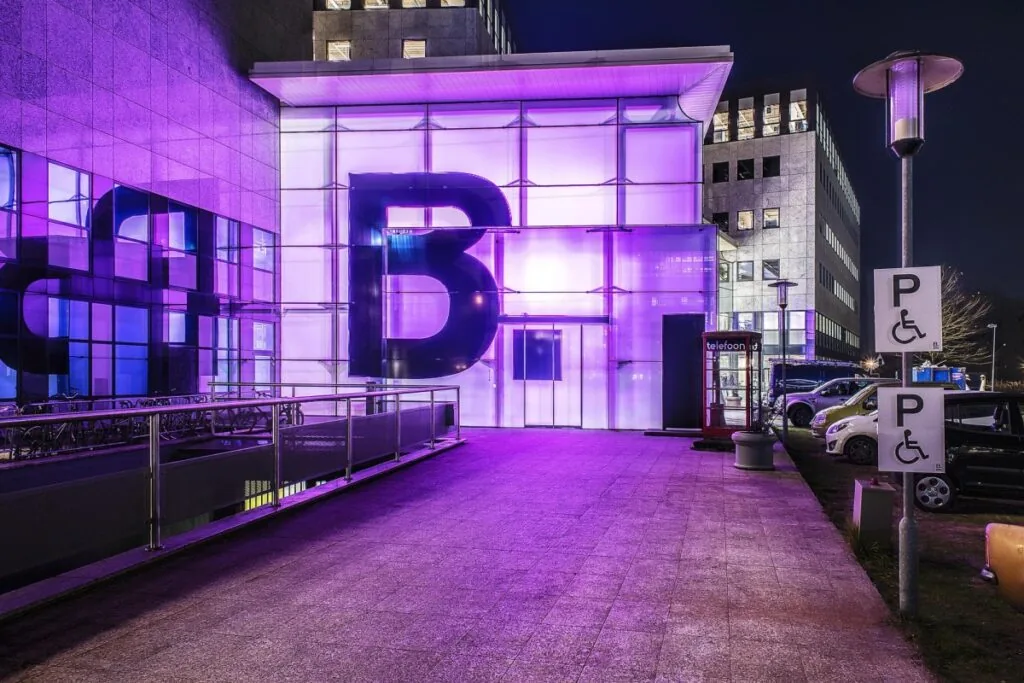 Entree van een modern kantoorgebouw aan het John M. Keynesplein in Amsterdam, verlicht met paarse verlichting en voorzien van grote letter B op de gevel.