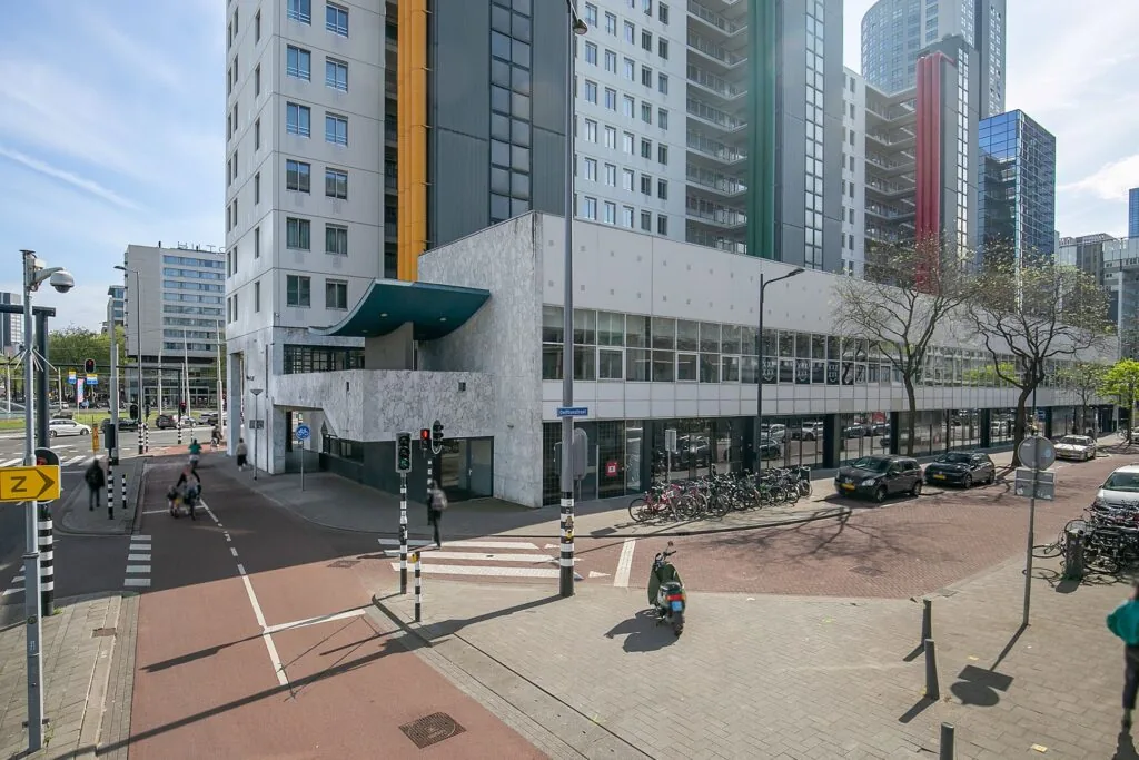 Straatbeeld van het gebouw Weena 113 in Rotterdam met omliggende hoogbouw, fietsenrekken en verkeerslichten op de kruising.