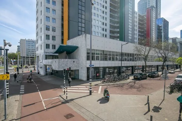 Straatbeeld van het gebouw Weena 113 in Rotterdam met omliggende hoogbouw, fietsenrekken en verkeerslichten op de kruising.