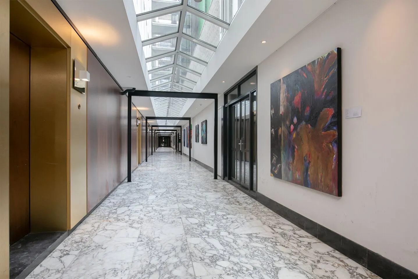 Lichte marmeren gang in Weena 81 met glazen dak, liften links en kunstwerken aan de muur rechts.