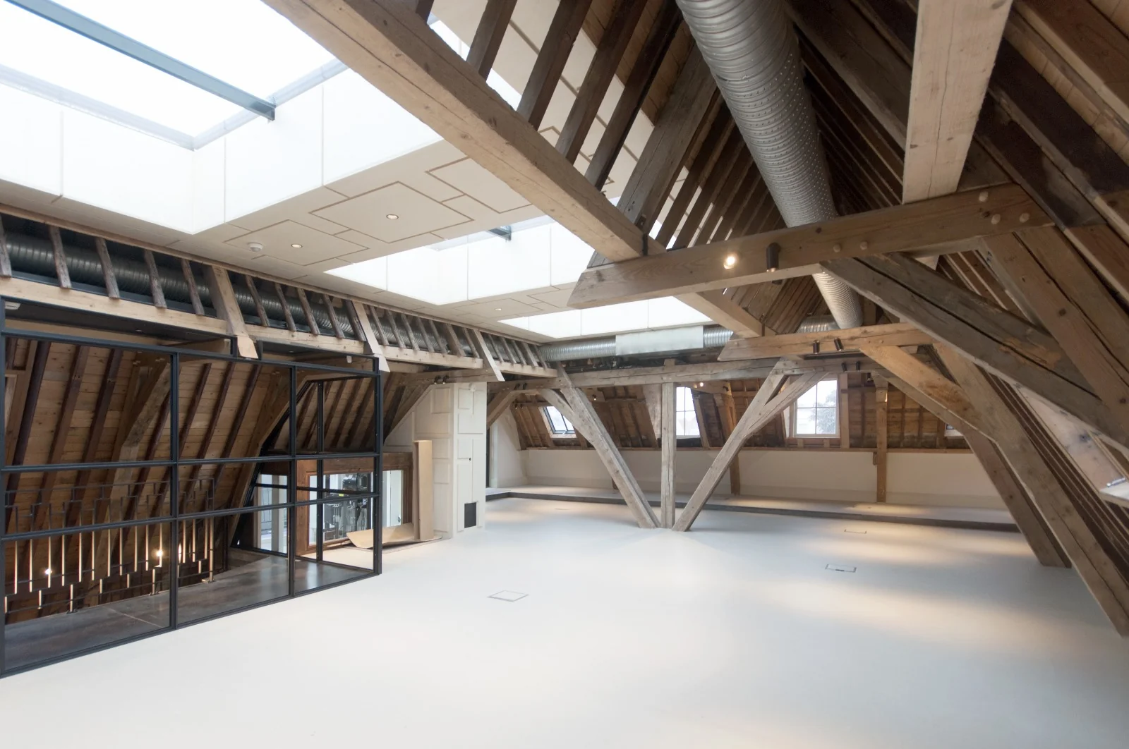 Moderne, gerenoveerde zolderruimte met zichtbare houten balken, grote dakramen en een strakke, witte vloer.